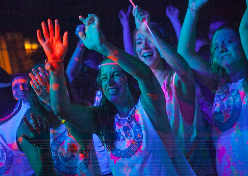 Excited crowd in neon-painted shirts dancing with hands raised at a GloZone college event