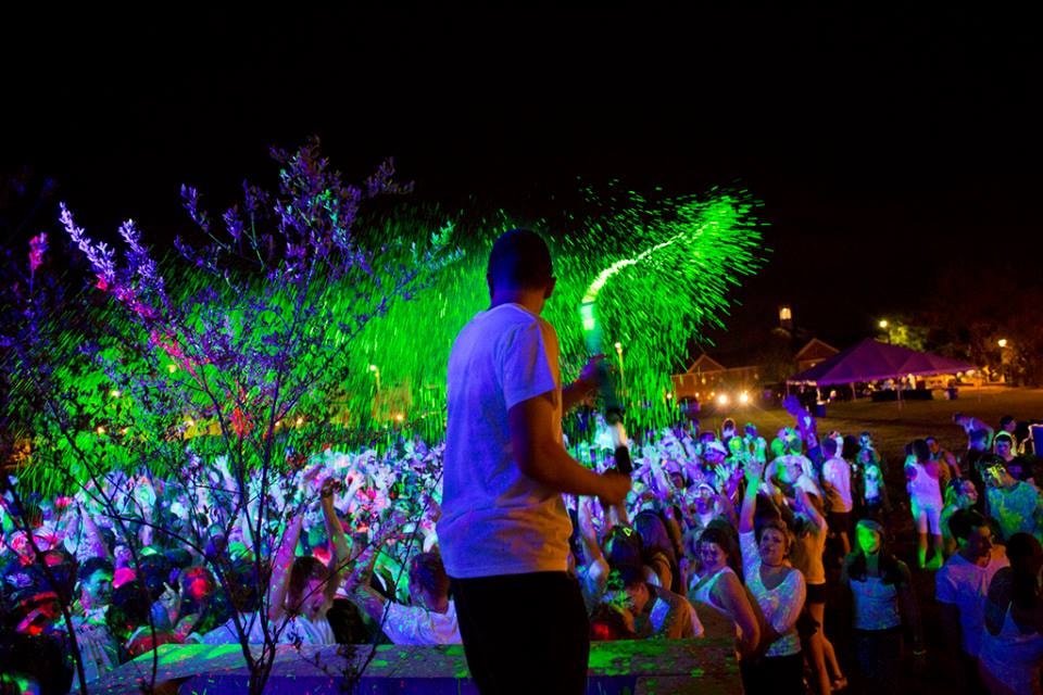DJ spraying neon green glow powder over a crowd surrounded by UV-lit trees at night